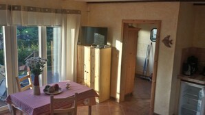 Una cocina pequeña con una mesa y sillas de madera, un televisor sobre un mueble de madera, y vistas al exterior a través de un ventanal.