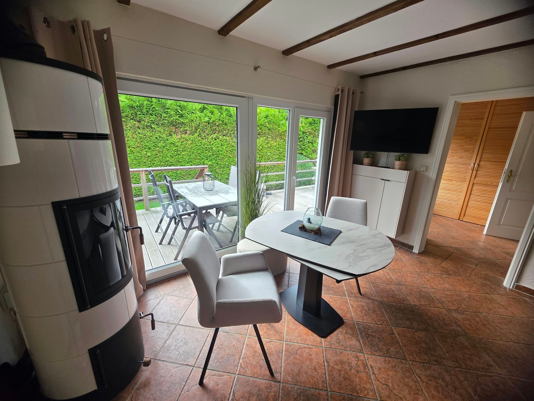 A modern kitchen with a dining area, a TV mounted above the cabinets, and a sliding glass door leading to a patio with a view of greenery.