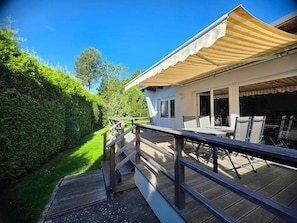 Una terraza de madera con mobiliario exterior y un porche cubierto, con vista a un jardín exuberante.