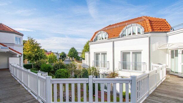 Ein Wohngebäude mit rotem Ziegeldach, weißen Außenwänden und einer Holzterrasse mit weißem Zaun.