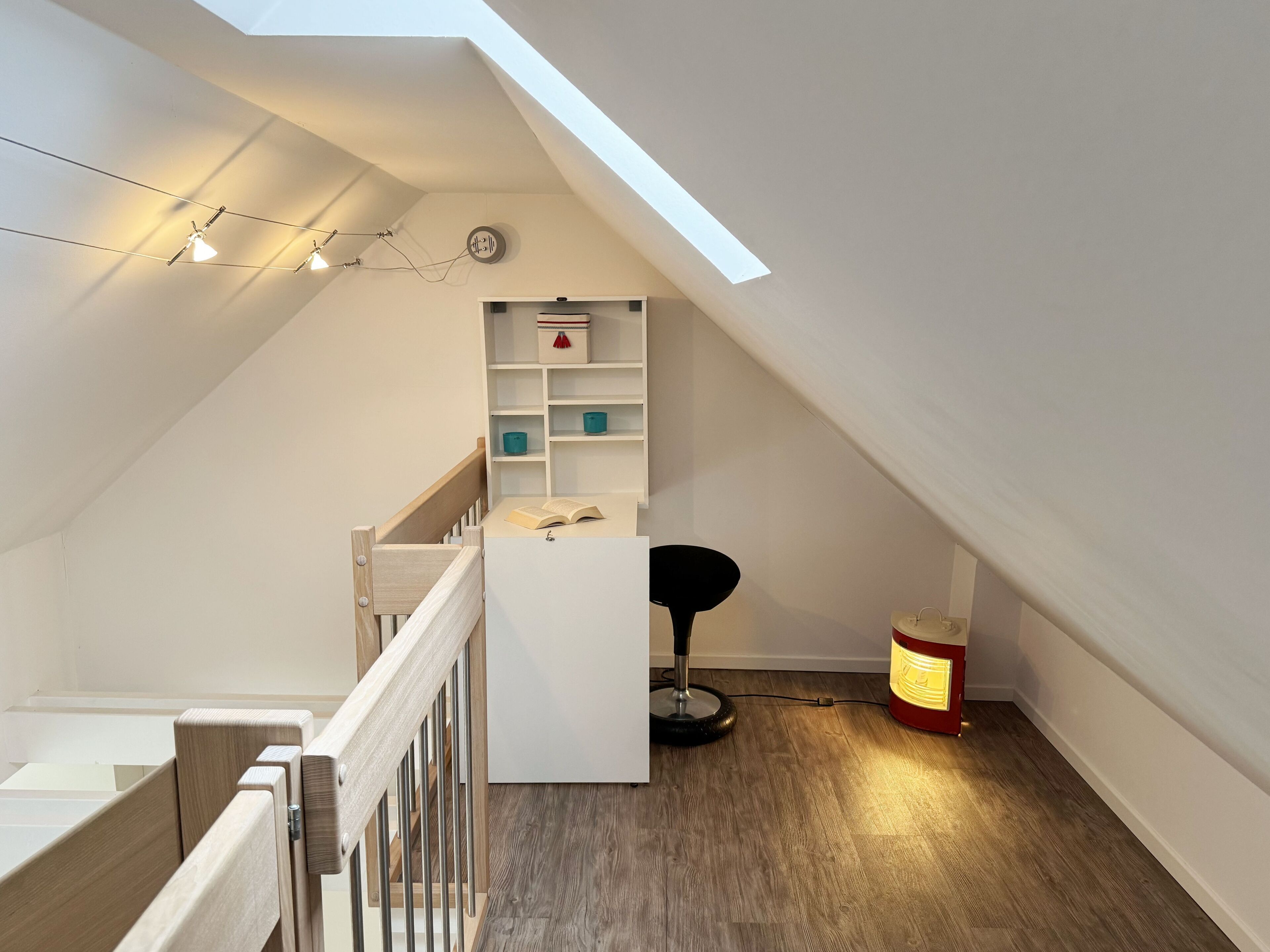 Ein modernes, helles Dachzimmer mit einem Oberlicht, einer Holztreppe, einem weißen Schreibtisch mit Stuhl und einem kleinen Tisch mit Lampe.