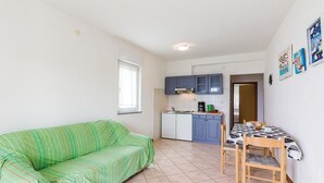 Ein kompakter Wohnraum mit Küchenzeile, einem grünen Sofa, einem Esstisch mit Stühlen und einem Fenster.