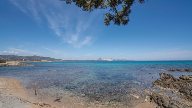 Una playa con aguas turquesas cristalinas, costa rocosa y montañas a lo lejos.