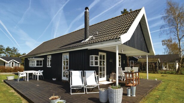 A black house with a white porch, a chimney, and a deck with outdoor furniture. - 2 bedroom amazing home in Væggerløse (Væggerløse)