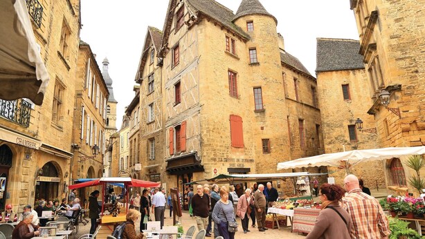 Un marché animé en plein air, situé sur une place historique bordée de bâtiments en pierre et dotée d’espaces de repos extérieurs.