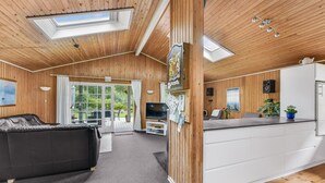 A modern cabin interior with a wooden ceiling, a living area with a sofa, a TV, and a kitchen with a fridge.