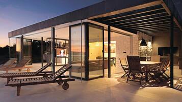 A modern outdoor patio with lounge chairs and a dining area, featuring glass walls and a stone wall.