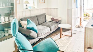 A living room with a grey sofa, a teal armchair, a small white table, and a glass cabinet with dishes. - Nice home in Süssau/Ostsee with sauna (Süssau/Ostsee)