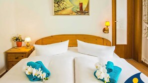 Hotelkamer met een houten hoofdeinde, twee witte bedden, een schilderij van een zeilboot op een klif, een nachtkastje met een lamp en een deur met gordijn.