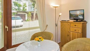 Een kamer met een glazen tafel, rieten stoelen, een televisie op een houten kast en uitzicht op de straat.