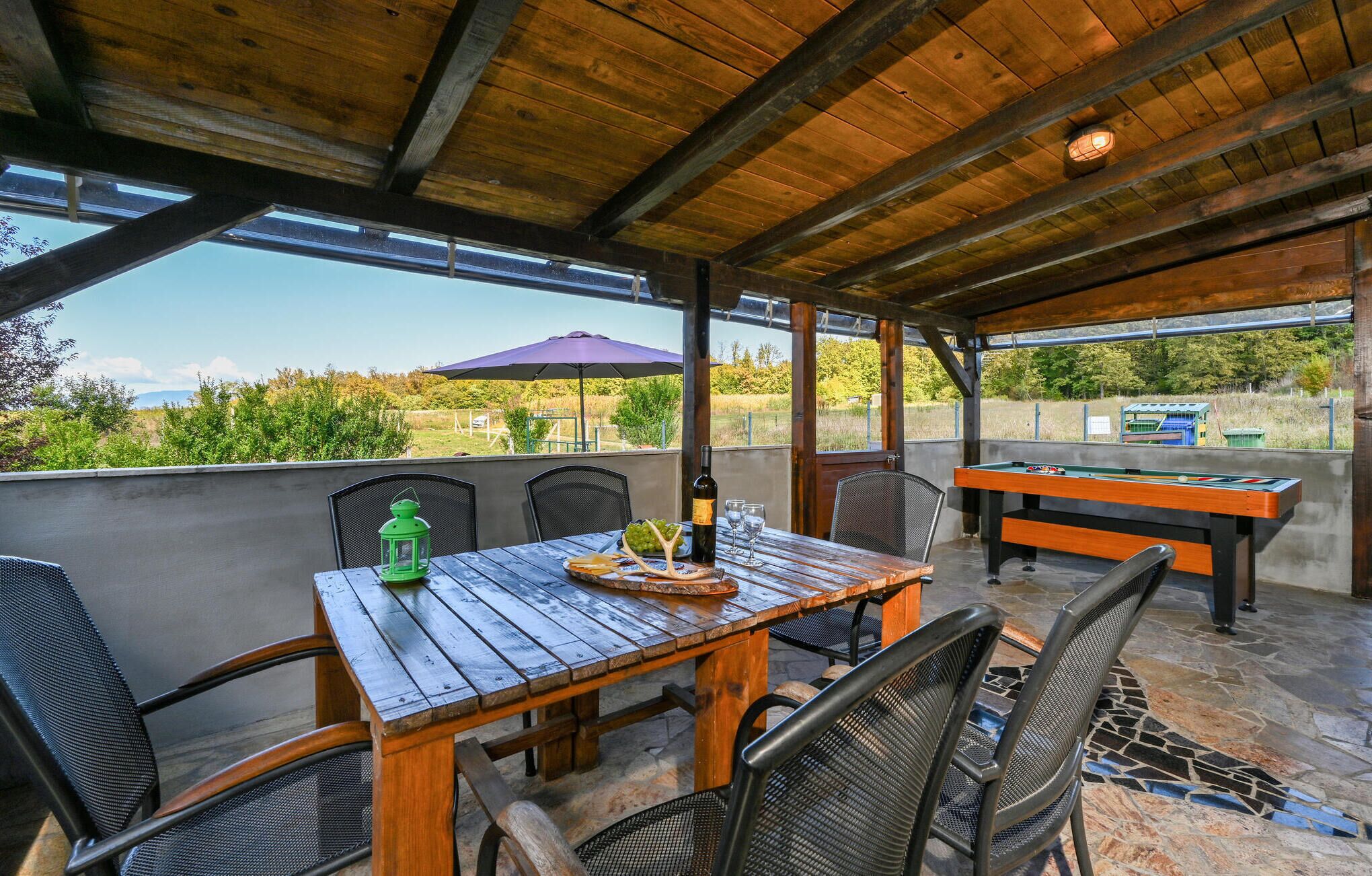 Um pátio externo com mesa de madeira, cadeiras, mesa de bilhar e vista para o campo.