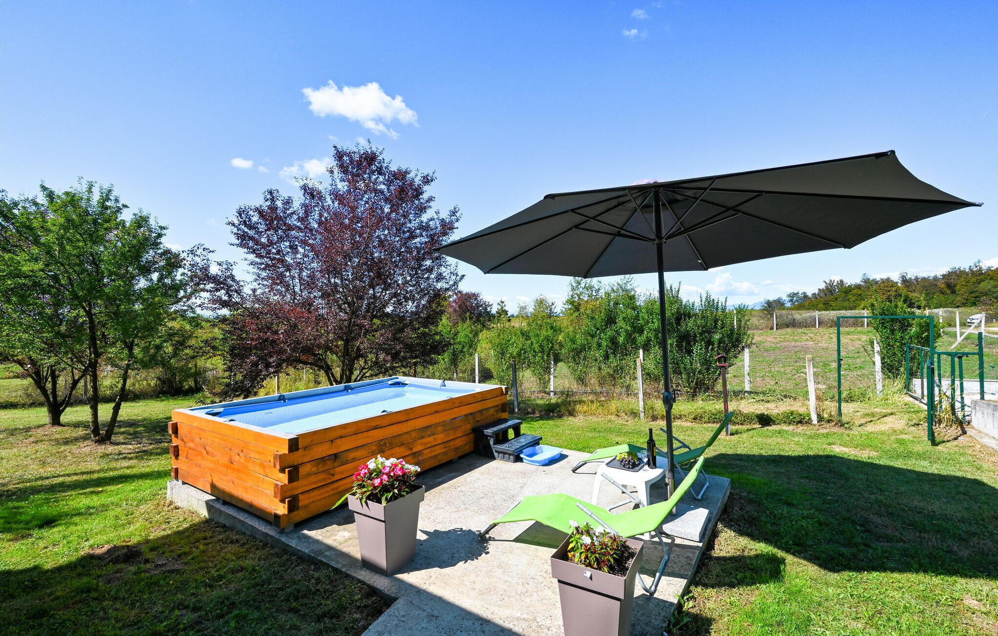 Een houten kuip waarin warm water staat, met een blauw binnenwerk, omgeven door een betonnen terras, een groene parasol en een bloempot.