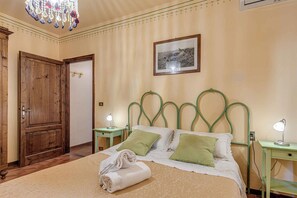 A bedroom with a large bed, a wooden wardrobe, a green metal headboard, a framed picture on the wall, and a chandelier.
