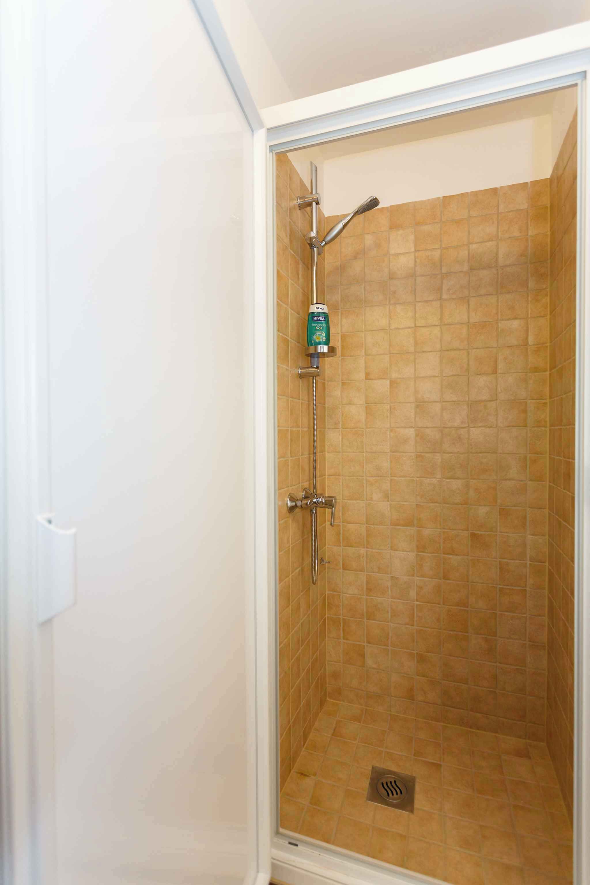 A glass-enclosed shower with beige tiles, a handheld showerhead, and a soap dispenser.