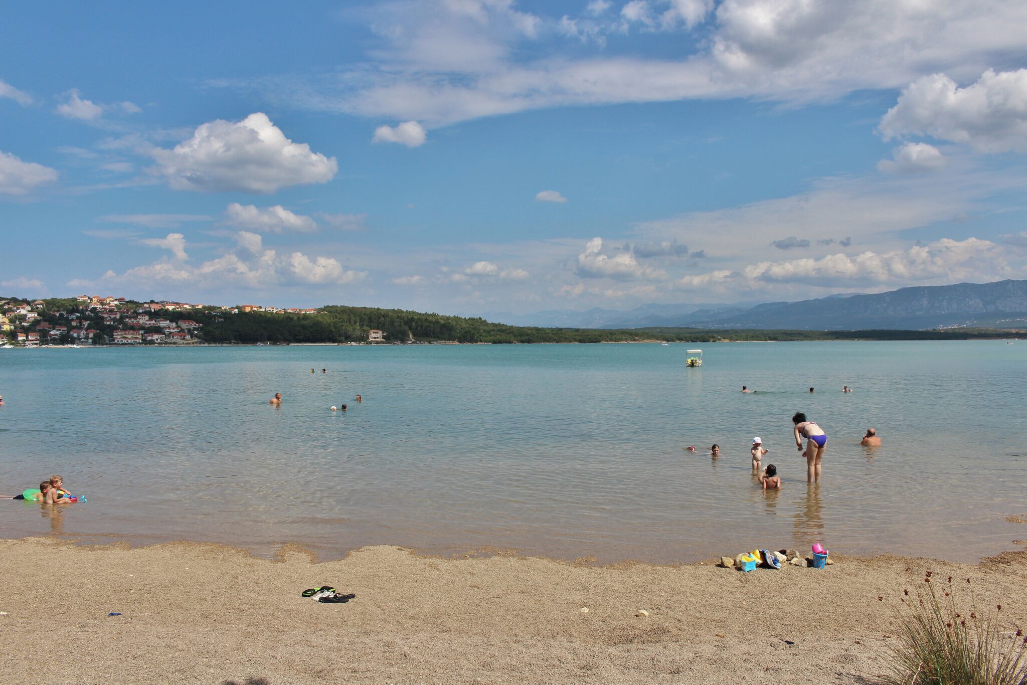 A beach with people swimming and sunbathing, a clear sky, and distant mountains.