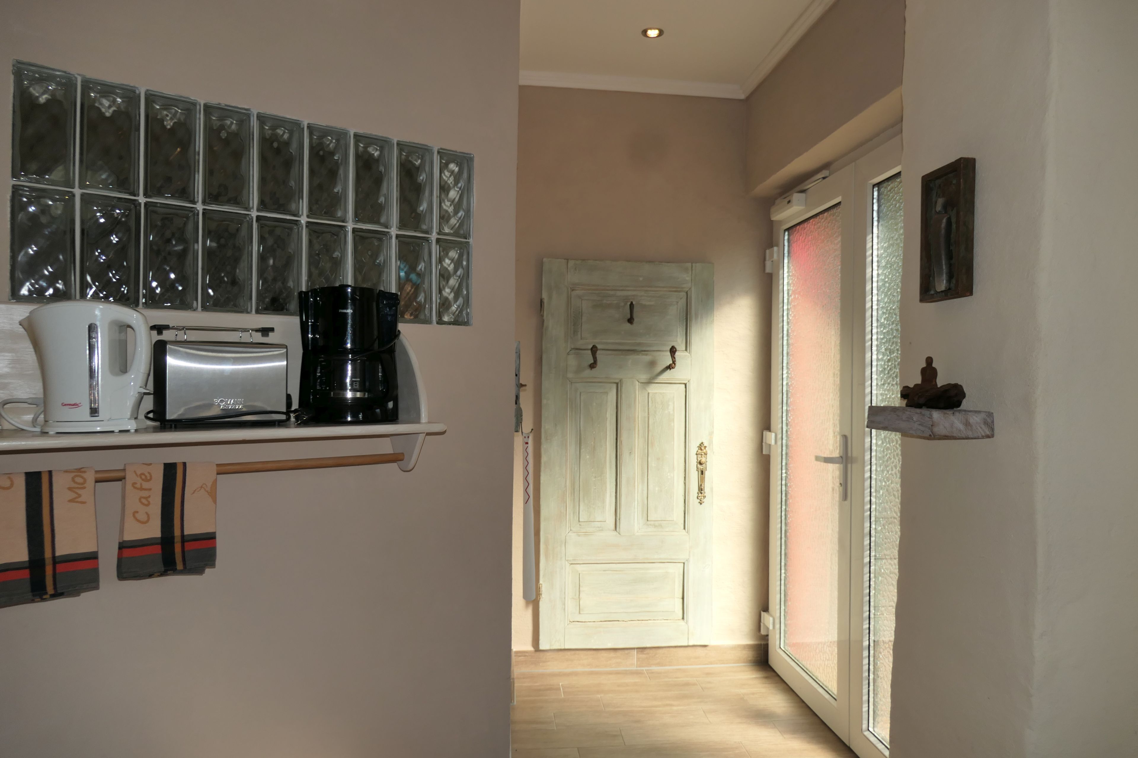 Una cocina con cafetera, tostadora y hervidor sobre una estantería, una puerta con panel de vidrio esmerilado y una pequeña repisa con un objeto decorativo.