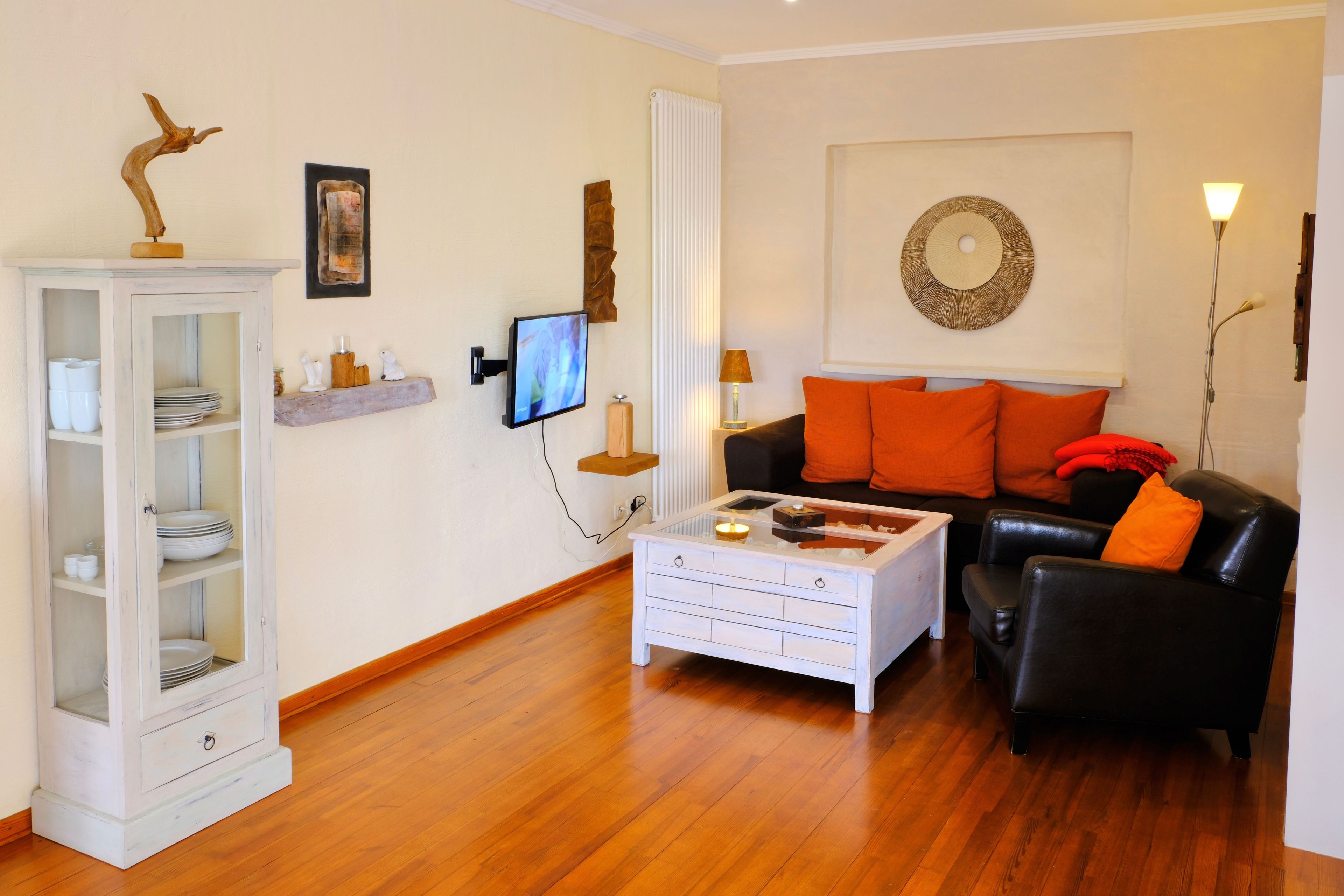 Una sala de estar con un mueble blanco, un sofá de cuero negro con cojines naranjas, una mesa de centro blanca y un televisor montado en la pared.