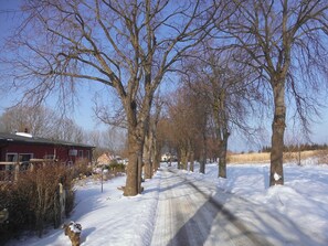 A snow-covered rural road flanked by leafless trees and a red building with a sign.