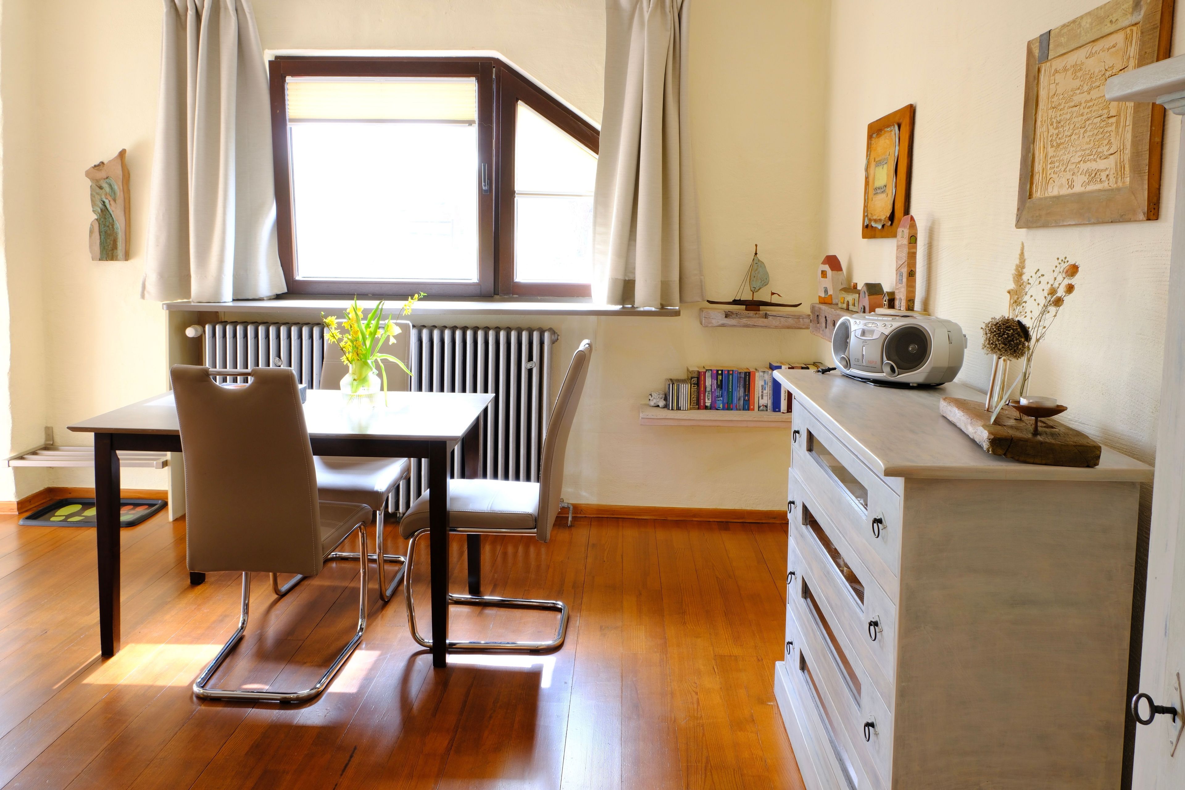 A dining area with a table, chairs, a radiator, a window with curtains, and a radio on a dresser.