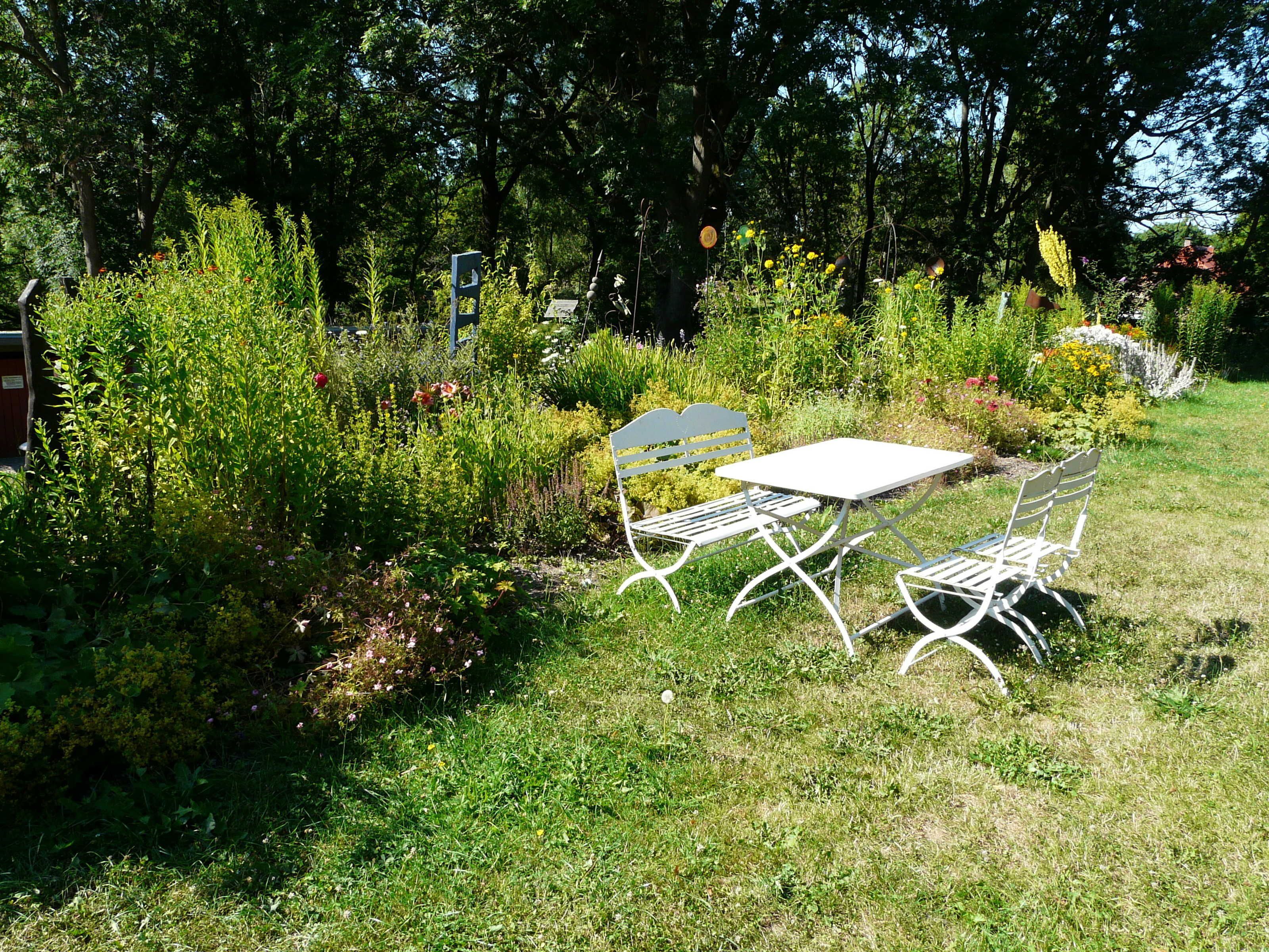 Un jardín con una mesa y sillas de metal blanco, rodeado de diversas plantas y árboles.