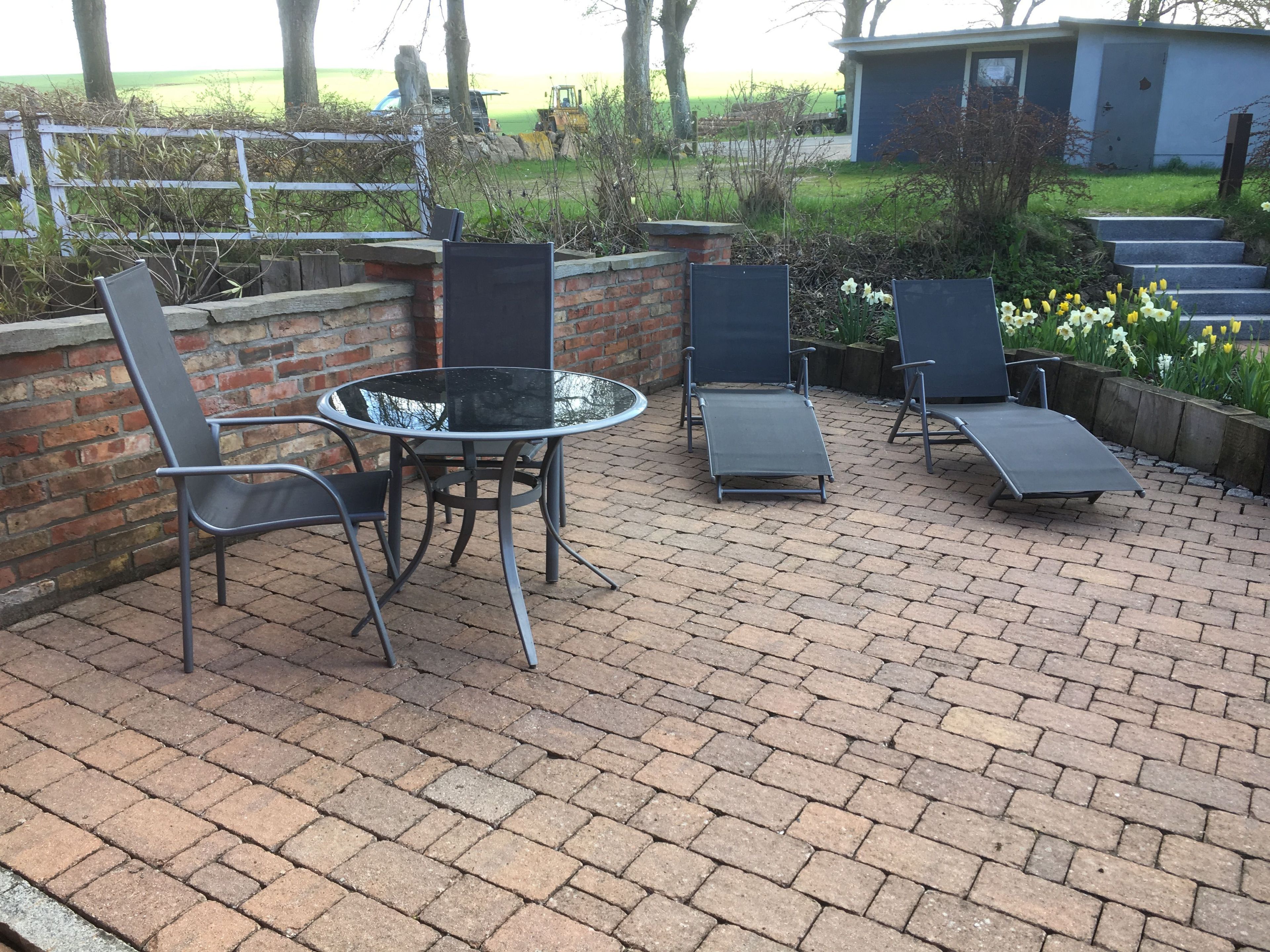 An outdoor patio with a round table and four chairs, a brick wall, and a garden with flowers.
