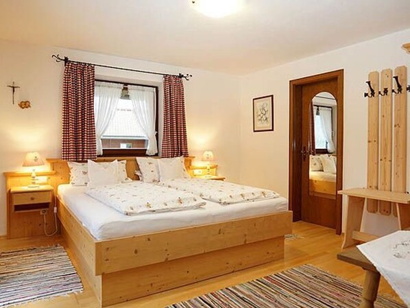 Un dormitorio con una cama de madera, dos mesitas de noche con lámparas, un ventanal con cortinas, un espejo y una silla de madera.