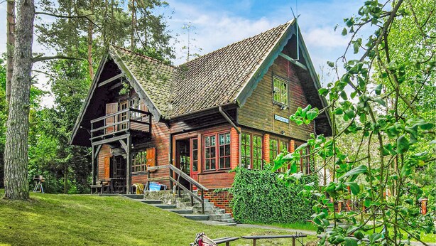 A rustic wooden house with a steeply pitched roof, a balcony, and a staircase leading to the upper floor.