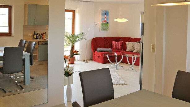 Un saló modern amb un sofà vermell, una taula de dinar amb caires i una cuina visible a través d'una porta oberta.