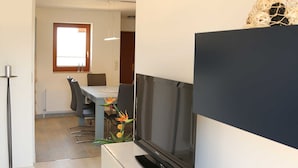 Un menjador modern amb un televisor de pantalla plana fixat a una paret blanca, una zona de dinar amb taula i cadires, i un passadís que condueix a una altra habitació.