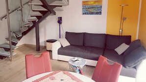 Uma sala de estar com um sofá cinza, uma poltrona vermelha, uma mesa de jantar com toalha quadriculada, uma escada de vidro e um quadro na parede.