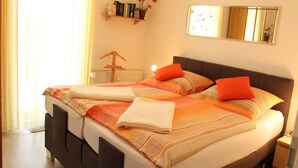 Ein ordentlich bezogenes Bett mit orange-weißer Bettwäsche, ein Nachttisch mit Lampe und ein wandmontierter Spiegel.