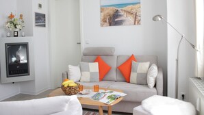Ein modernes Wohnzimmer mit einem grauen Sofa, orangefarbenen Kissen, einem Couchtisch aus Holz mit Obstschale und Zeitschrift, einem weißen Sessel und einem Bild von einem Strandweg an der Wand.