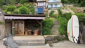 Eine Holzterrasse mit überdachter Sitzgruppe, einer Steintreppe und einer Reihe Surfbretter.