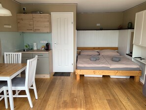 Een knus kamertje met een houten bed, een klein bureau, een stoel en een kitchenette.