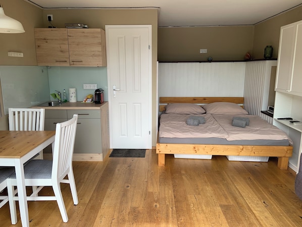 Una habitación compacta con una cama de madera, un escritorio pequeño, una silla y una zona de kitchenette.