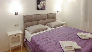 Una cama bien hecha con un edredón morado, dos almohadas y toallas dobladas sobre una mesita de noche blanca.