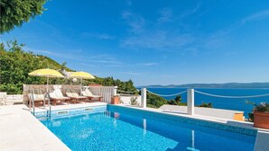 Una piscina con sillas de descanso y sombrillas, rodeada por un muro de piedra y con vistas al litoral.