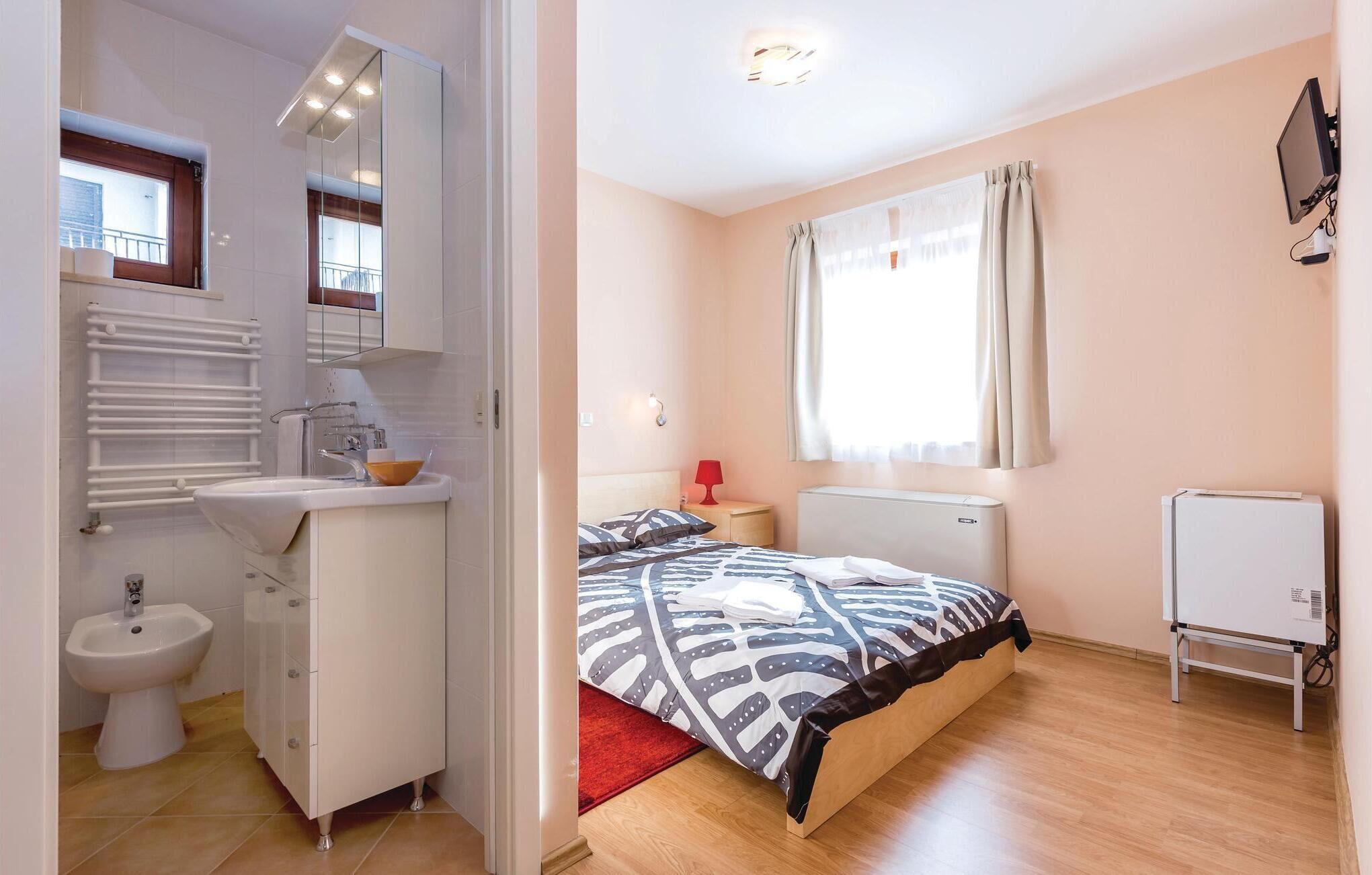 Un dormitorio con cama, mesita de noche, radiador, ventana con cortinas y televisor montado en la pared, además de un baño con lavamanos, bidé y toallero.