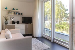 A modern living room with a sofa, a flat-screen TV, a bookshelf, and a large window with a view of trees.