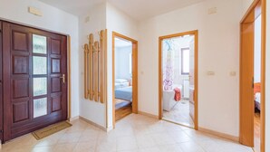 Un couloir avec une porte en bois, un porte-manteau et, au fond, on aperçoit une chambre avec un lit et une fenêtre.