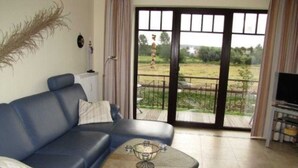 A living room with a blue sofa, a coffee table, and a view of a garden through a sliding glass door. - (257) 3-room apartment - apartment complex holiday dreams (Kühlungsborn)