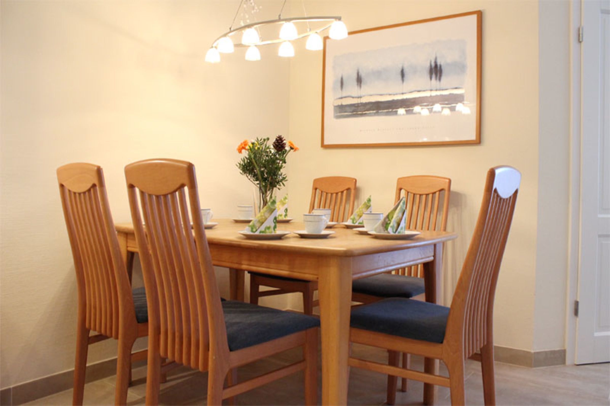 A dining area with a wooden table and chairs, a vase with flowers, and a framed picture on the wall.