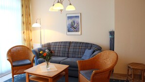 Una sala de estar con un sofá, dos sillones de mimbre, una mesa de centro con un jarrón de flores, una lámpara de pie y un cuadro en la pared.
