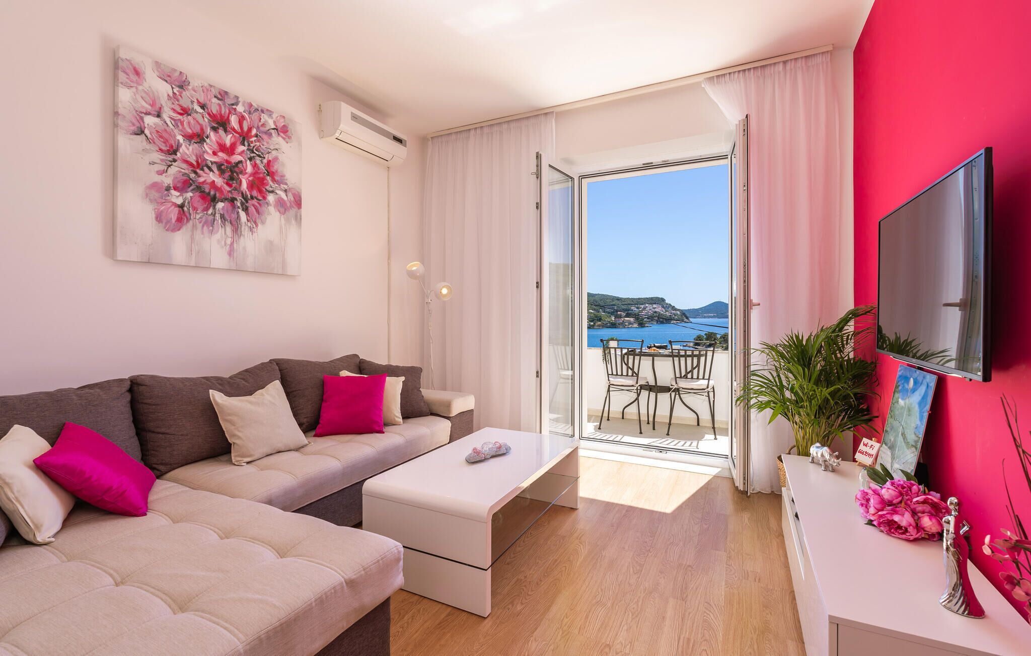 Una sala de estar moderna con un sofá, una mesa de centro y un televisor sobre un mueble. Hay un balcón con vista al mar y una mesa con sillas.