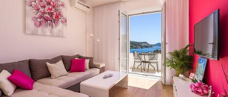 Una sala de estar moderna con un sofá, una mesa de centro y un televisor sobre un mueble. Hay un balcón con vista al mar y una mesa con sillas.