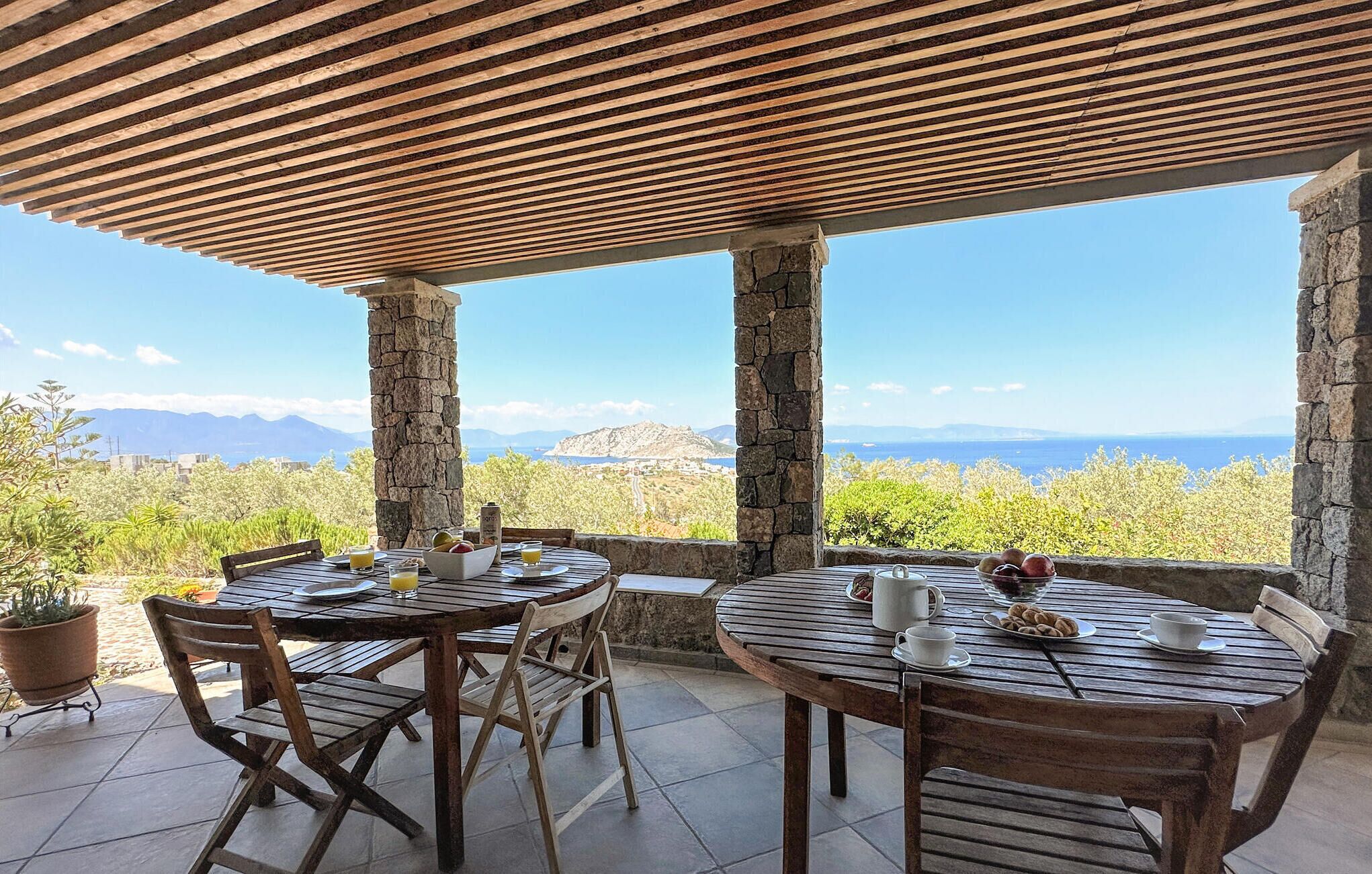 A terrace with wooden tables and chairs, stone pillars, and a view of mountains and the sea.