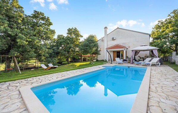Ein Swimmingpool mit Liegestühlen und einem überdachten Sitzbereich, umgeben von einer Steinterrasse und üppiger Bepflanzung.