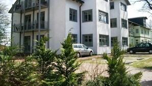 Un immeuble à plusieurs étages, doté d’une façade blanche, de nombreux balcons et d’un espace de stationnement où se trouvent deux voitures.