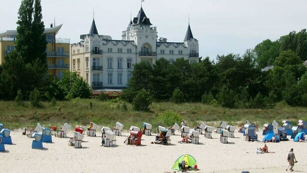 Une plage avec des chaises longues et des tentes, un château en arrière-plan, et une personne qui marche sur le sable.