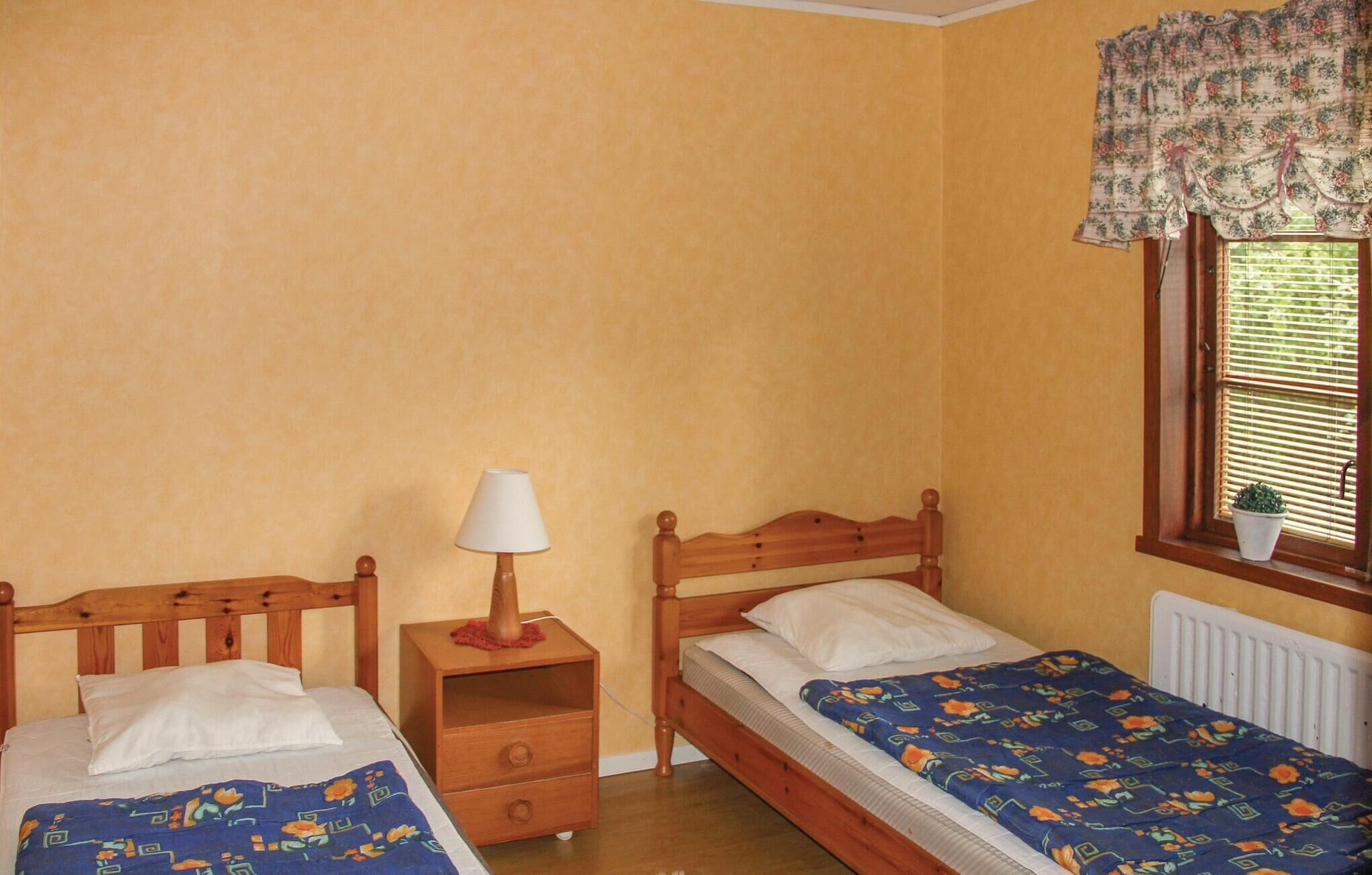 A bedroom with two single beds, a nightstand with a lamp, a window with blinds, and a potted plant.